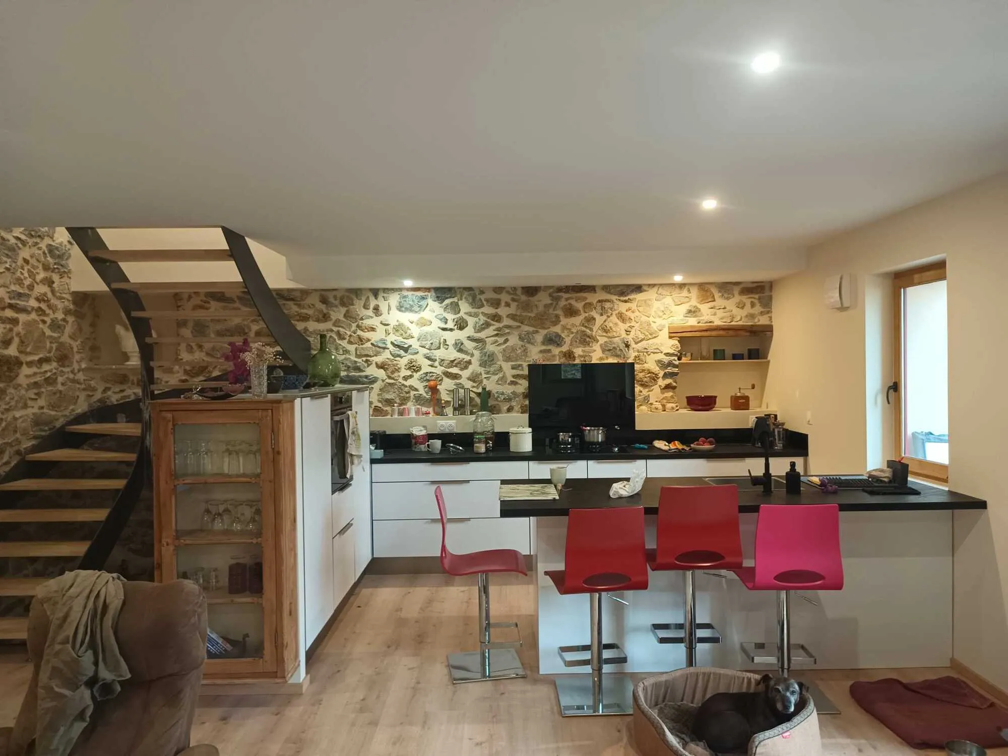 Cuisine ouverte avec îlot central, mur en pierre apparente et escalier en bois.
